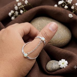 May include: A silver double-band ring connected by a delicate silver chain. Each band features a small, round, iridescent opal stone set in a simple bezel. The two bands are designed to be worn on the thumb and index finger simultaneously.