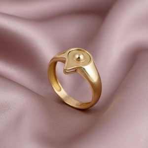 May include: A gold-colored ring with a unique design. The ring features a rounded band and a top with a teardrop shape and a small, raised sphere. The ring is set against a pink fabric background.