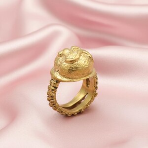 May include: A gold-colored ring with a textured design. The ring features a large, textured top with a rounded shape and a band with small, round details. The ring is set against a pink satin background.