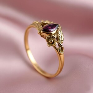 Puede incluir: Anillo dorado con una gema púrpura en forma de marquesa. El anillo presenta detalles florales y de hojas. El fondo es de color rosa pálido.