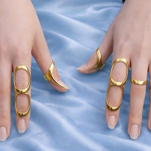 Può includere: Anelli dorati in vari design. Due dita presentano anelli ovali allungati, mentre altri hanno fasce curve. Gli anelli sono esposti su un tessuto blu chiaro.