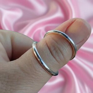 May include: Two silver rings are worn on a finger. The rings are thin and have a smooth, shiny surface. The background is a soft pink color.