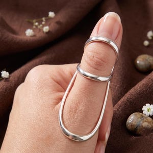 May include: A silver-coloured metal finger splint featuring a smooth, polished finish. The device consists of two circular bands connected by two parallel vertical bars that extend along the length of the finger to provide support and stabilisation.