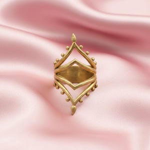 Puede incluir: Un anillo dorado con un diseño intrincado. El anillo presenta un elemento central en forma de diamante, enmarcado por formas geométricas y pequeñas esferas decorativas. El anillo está sobre un fondo rosa suave.