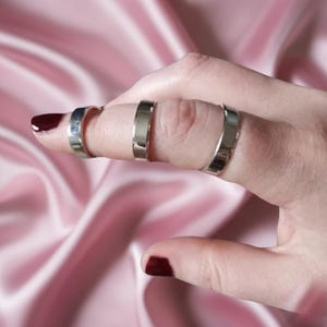 May include: A hand with three silver rings worn on the index finger. The rings are simple bands of varying widths. The fingernails are painted with a dark red nail polish. The background is a soft, pink fabric.