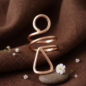 May include: A rose gold-toned ring featuring a continuous wire design that forms a circular loop at the top and a triangular shape at the bottom. The metal band wraps around itself, creating an open, geometric silhouette with a polished finish.