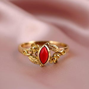 May include: A gold ring featuring a marquise-cut red gemstone. The ring has a floral and leaf design on the band, with small flowers and leaves on either side of the gemstone. The ring is set against a soft pink background.