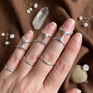Könnte beinhalten: Ein Set aus fünf silbernen Ringen an den Fingern. Drei der Ringe haben ein markantes H-förmiges Design, das über das Fingergelenk reicht, während zwei schlichte, glatte und polierte Silberringe an der Fingerbasis getragen werden.