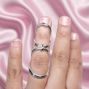 Puede incluir: Una férula de dedo plateada que se usa en un dedo. La férula tiene tres bandas conectadas que envuelven el dedo. El fondo es una tela rosa y sedosa.