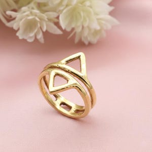 Op de afbeelding: Een goudkleurige ring met een dubbel bandontwerp. De ring heeft twee open driehoeken, de ene boven de andere, waardoor een geometrisch patroon ontstaat. De ring staat tegen een zachtroze achtergrond met witte bloemen.