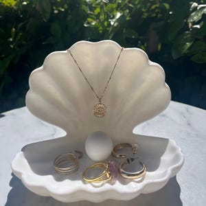 Puede incluir: Un joyero blanco en forma de concha contiene un collar de oro con un colgante de sol, un huevo blanco y varios anillos y pendientes de oro. El joyero está sobre una superficie blanca, con un fondo verde borroso.