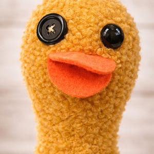 Puede incluir: Un juguete de pato de peluche hecho a mano con un cuerpo texturizado de color amarillo dorado. Cuenta con un ojo de botón negro, un ojo de botón negro con un diseño de cruz y un pico de fieltro naranja. El juguete está diseñado para parecerse a un pato.