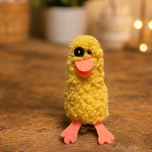 May include: A small, yellow, textured plush duck figure with a fuzzy, curly surface. It features a single black button eye, a flat orange felt beak, and two orange felt webbed feet extending from the base of its cylindrical, upright body.