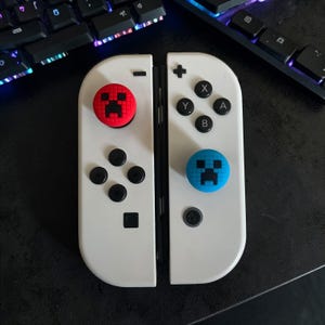 May include: A pair of white game controllers featuring black buttons and directional pads. The left controller has a red thumbstick cover with a black pixelated face, and the right controller has a blue thumbstick cover with a black pixelated face.