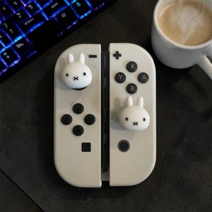 May include: A pair of off-white video game controllers featuring two white rabbit-shaped joystick covers. The controllers include black buttons labelled with X, Y, A, and B, a plus sign, a minus sign, and directional buttons on a smooth, matte surface.