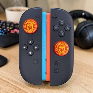May include: A pair of black game controller attachments featuring the Manchester United logo in red and yellow on each side. The controllers have standard buttons, a directional pad, and light blue and orange side accents for a gaming console.