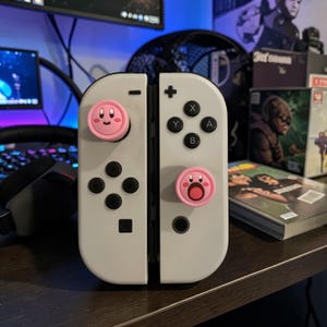 May include: A pair of white video game controllers featuring two pink circular thumbstick covers. The left cover displays a smiling face, while the right cover shows an open-mouthed surprised expression. The controllers include black buttons.