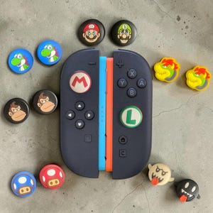 May include: A black game controller with a blue and orange side rail, featuring a red M thumbstick cover and a green L thumbstick cover. Surrounding the controller are various circular thumbstick covers with character graphics including Mario, Luigi, Yoshi, Donkey Kong, Bowser, a blue mushroom, a red mushroom, a Boo ghost, and a Bullet Bill.