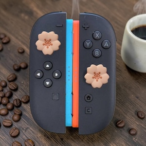 May include: A pair of black game controllers featuring light blue and orange side rails, with two pale pink cherry blossom-shaped thumbstick covers attached to the joysticks. The controllers include standard buttons labelled with A, B, X, Y, plus, minus, home, and capture icons.