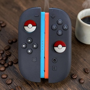 May include: A pair of black video game controllers featuring custom joystick caps designed to look like red and white Poké Balls. The controllers are joined together, displaying a light blue side edge and an orange side edge with directional buttons.
