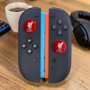 May include: A pair of black game controllers featuring a light blue and orange side panel. Each controller has a circular red emblem with a white Liver bird logo and the text L.F.C. printed on the front. The controllers include standard buttons.