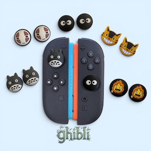 May include: A dark grey video game controller with a blue and orange side rail, featuring various circular silicone thumbstick covers with character designs including soot sprites, spirits, cats, and fire creatures, with the text "ghibli" below.
