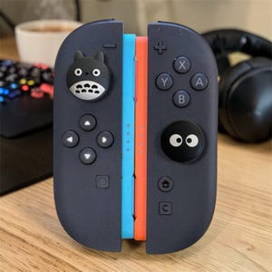 May include: A pair of dark grey gaming controllers attached together, featuring a light blue side and an orange side. The left controller has a black Totoro-shaped thumbstick cover, and the right controller has a black circular thumbstick cover with two white eyes.