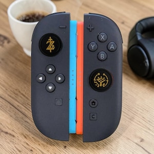 May include: A pair of black game controllers featuring a light blue and orange side edge. The left controller displays a gold Z symbol on the joystick, while the right controller features a gold eye icon on the joystick and standard A, B, X, Y buttons.