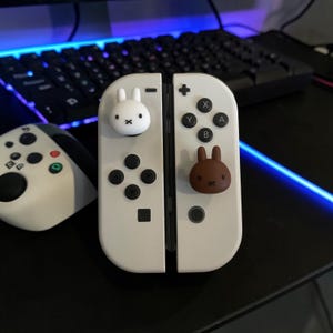May include: A pair of white game controllers featuring two small, three-dimensional rabbit-shaped thumbstick covers. One cover is white with a black cross-stitch mouth and eyes, while the other is brown with identical facial features.