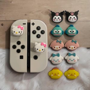 May include: A pair of off-white game controllers with two Hello Kitty character thumb grip covers attached. Beside them are ten additional silicone character thumb grip covers featuring various designs including Kuromi, Hangyodon, My Melody, Cinnamoroll, and Pompompurin.