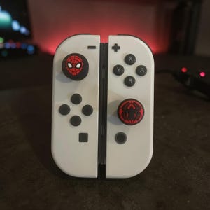 May include: A pair of white game controllers featuring black thumbstick covers. The left thumbstick cover displays a red and black Spider-Man mask graphic, while the right thumbstick cover features a red and black spider emblem design.