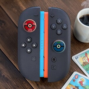 May include: A pair of black game controllers featuring a light blue side rail and an orange side rail. Each controller has a circular thumbstick cover, one red with a white centre and one blue with a green centre, both resembling spherical containers.