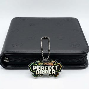 May include: A black zipped binder featuring an embossed pattern of circular symbols across its surface. A silver-toned ball chain is attached to the zip pull, holding a clear acrylic charm with the text MEGA EVOLUTION PERFECT ORDER.