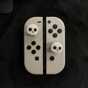 May include: A pair of off-white game controllers featuring two white skull-shaped joystick covers. The left controller has a minus button and a directional pad, while the right controller has a plus button, four action buttons labelled A, B, X, Y.