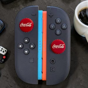 May include: A pair of black game controllers featuring a light blue and orange side rail. Each controller is adorned with a circular red Coca-Cola bottle cap emblem. The controllers include standard directional buttons, action buttons, and icons.