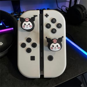 May include: A pair of white gaming controllers featuring black and pink Kuromi character thumbstick covers. The left controller has a directional pad and a thumbstick, while the right controller has A, B, X, Y buttons and a thumbstick.