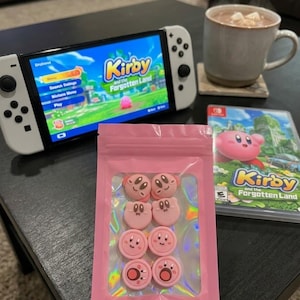 May include: A set of eight pink silicone thumbstick caps for a game controller, each featuring a different Kirby character expression, packaged in a small pink resealable bag with a clear window displaying the circular covers inside.