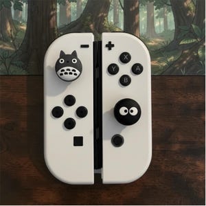 May include: A pair of white game controllers featuring black buttons and two custom joystick covers. One cover is shaped like a grey and white creature with ears, while the other is a black circular cap with two white eyes printed on the surface.