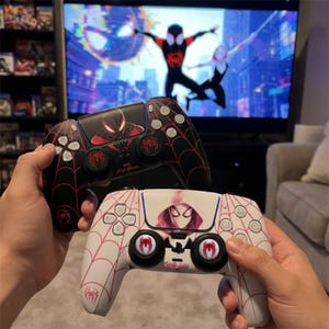 May include: Two wireless game controllers featuring Spider-Man themed designs. The top controller is black with a red spider web pattern and a Miles Morales graphic. The bottom controller is white with a red web pattern and a Spider-Gwen graphic.