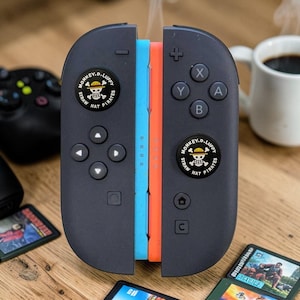 May include: A pair of dark grey game controller attachments featuring a circular black emblem with the white skull and crossbones logo of the Straw Hat Pirates and the text Monkey D. Luffy. The controllers have light blue and orange inner edges.