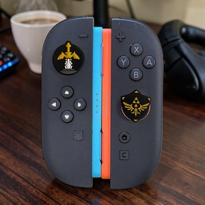 May include: Two black game controllers with a light blue and orange side edge. The left controller features a gold sword emblem on the joystick, while the right controller has a gold shield emblem on the joystick, both with directional buttons.