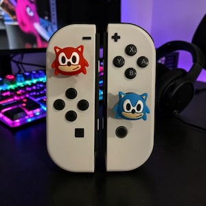 May include: A pair of white game controllers featuring two silicone thumbstick covers. One cover is a red character with pointed ears, and the other is a blue character with pointed ears. Both controllers have black buttons, a plus sign, and a minus sign.