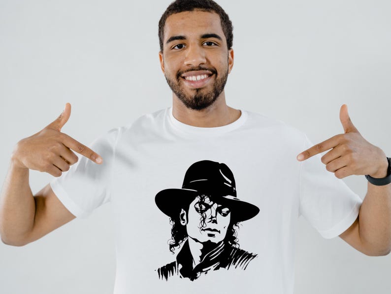 May include: White t-shirt featuring a black and white illustration of Michael Jackson wearing a hat. The image is a stylized portrait. The person in the image is pointing at the shirt.
