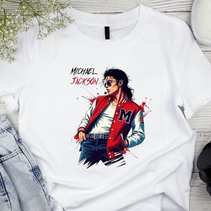May include: White t-shirt featuring a stylized illustration of Michael Jackson in a red and white jacket with the letter "M". The text "MICHAEL JACKSON" is above the illustration in red and black.