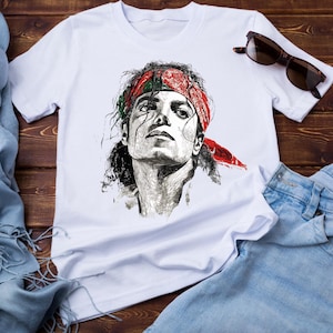 May include: White t-shirt featuring a detailed black and white portrait of a person wearing a red bandana. The shirt is paired with blue jeans and sunglasses, set against a wooden background.
