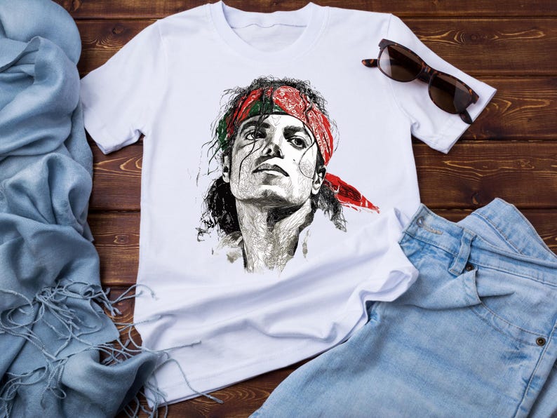 May include: White t-shirt featuring a detailed black and white portrait of a person wearing a red bandana. The shirt is paired with blue jeans and sunglasses, set against a wooden background.