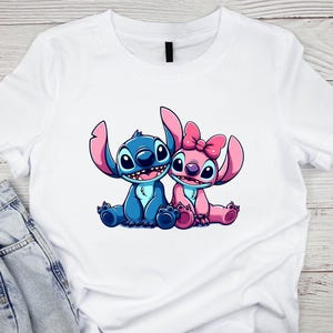 K&ouml;nnte beinhalten: Wei&szlig;es T-Shirt mit einer Cartoon-Illustration von zwei Figuren, eine blau und eine rosa, mit gro&szlig;en Ohren und l&auml;chelnden Gesichtern. Die rosa Figur hat eine Schleife. Das Design ist auf dem T-Shirt zentriert.