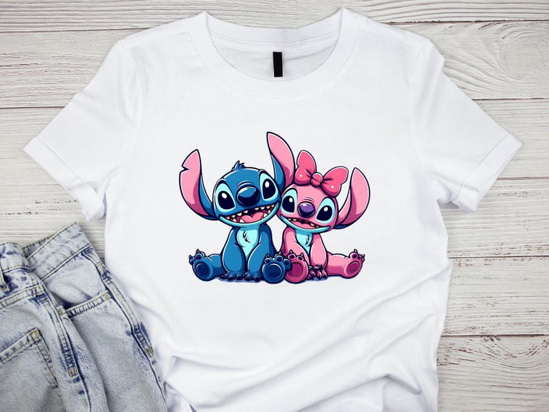 K&ouml;nnte beinhalten: Wei&szlig;es T-Shirt mit einer Cartoon-Illustration von zwei Figuren, eine blau und eine rosa, mit gro&szlig;en Ohren und l&auml;chelnden Gesichtern. Die rosa Figur hat eine Schleife. Das Design ist auf dem T-Shirt zentriert.