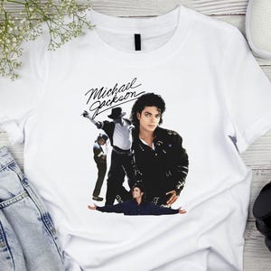 May include: White t-shirt featuring a graphic of Michael Jackson in various poses. The design includes the name "Michael Jackson" in a stylized script. The image shows the singer in a black jacket and hat.