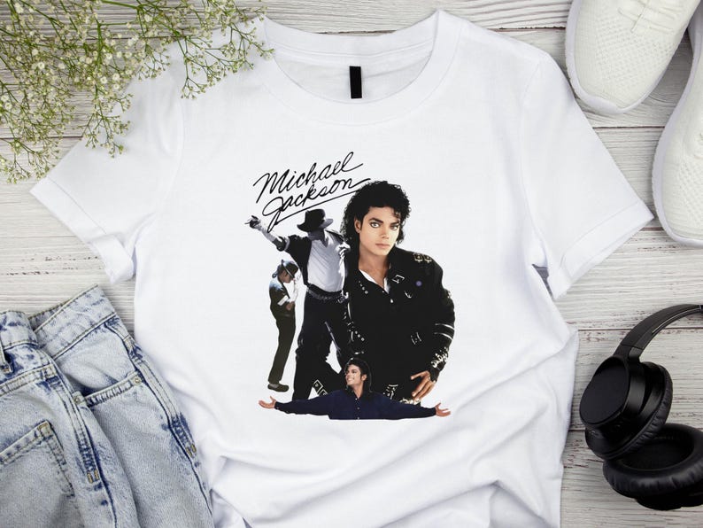 May include: White t-shirt featuring a graphic of Michael Jackson in various poses. The design includes the name "Michael Jackson" in a stylized script. The image shows the singer in a black jacket and hat.